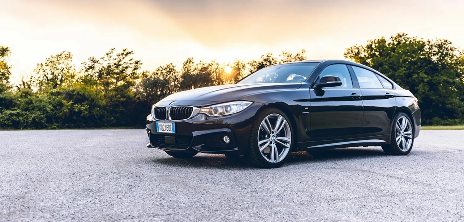 A black sporty BMW sedan is parked on an empty paved surface with trees and a sunset in the background.